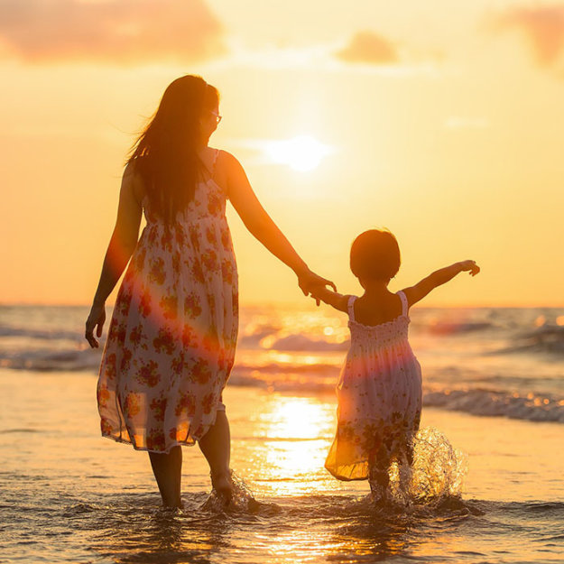 Mother and daughter holding hands wading in ocean at sunset.