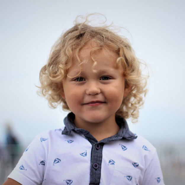 Young child with curly blonde hair smiling outdoors in purple patterned shirt.