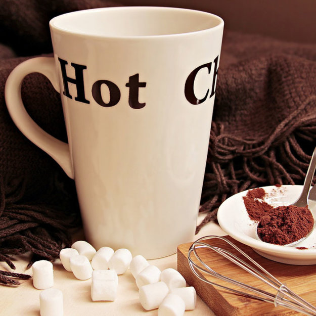 White mug with "Hot C" text surrounded by marshmallows, chocolate, and cozy blanket setup.