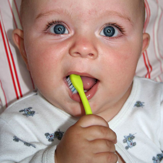 kid-toothbrush-2022_700.jpg Infant with blue eyes holding yellow toothbrush in mouth, wearing white patterned shirt.