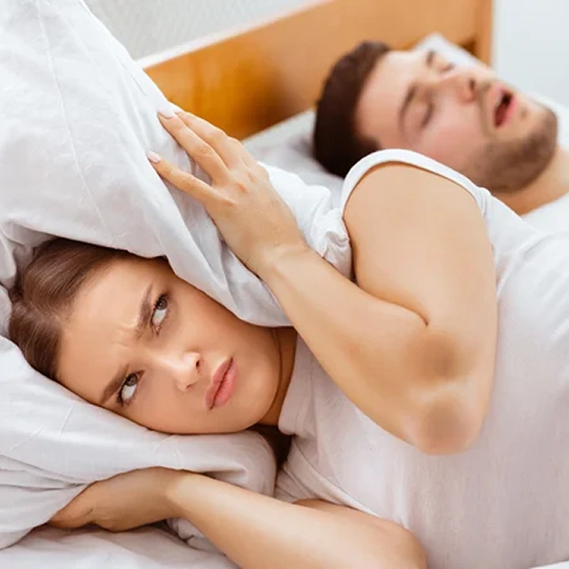 Woman covering her ears with a pillow while her partner snores beside her representing sleep apnea symptoms and dental treatment at Dentistry of Old Town Scottsdale