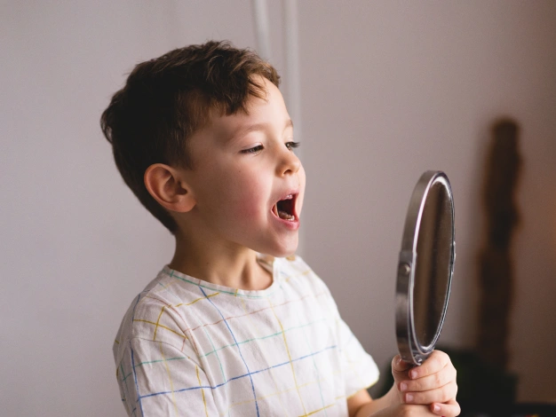 Child examining tongue movement in a mirror as part of a lingual frenectomy assessment for tongue tie treatment.
